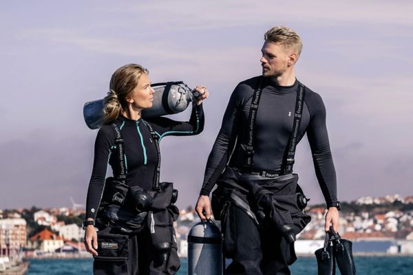 Two people wearing Fourth element black wetsuits with visible branding against a light purple sky.