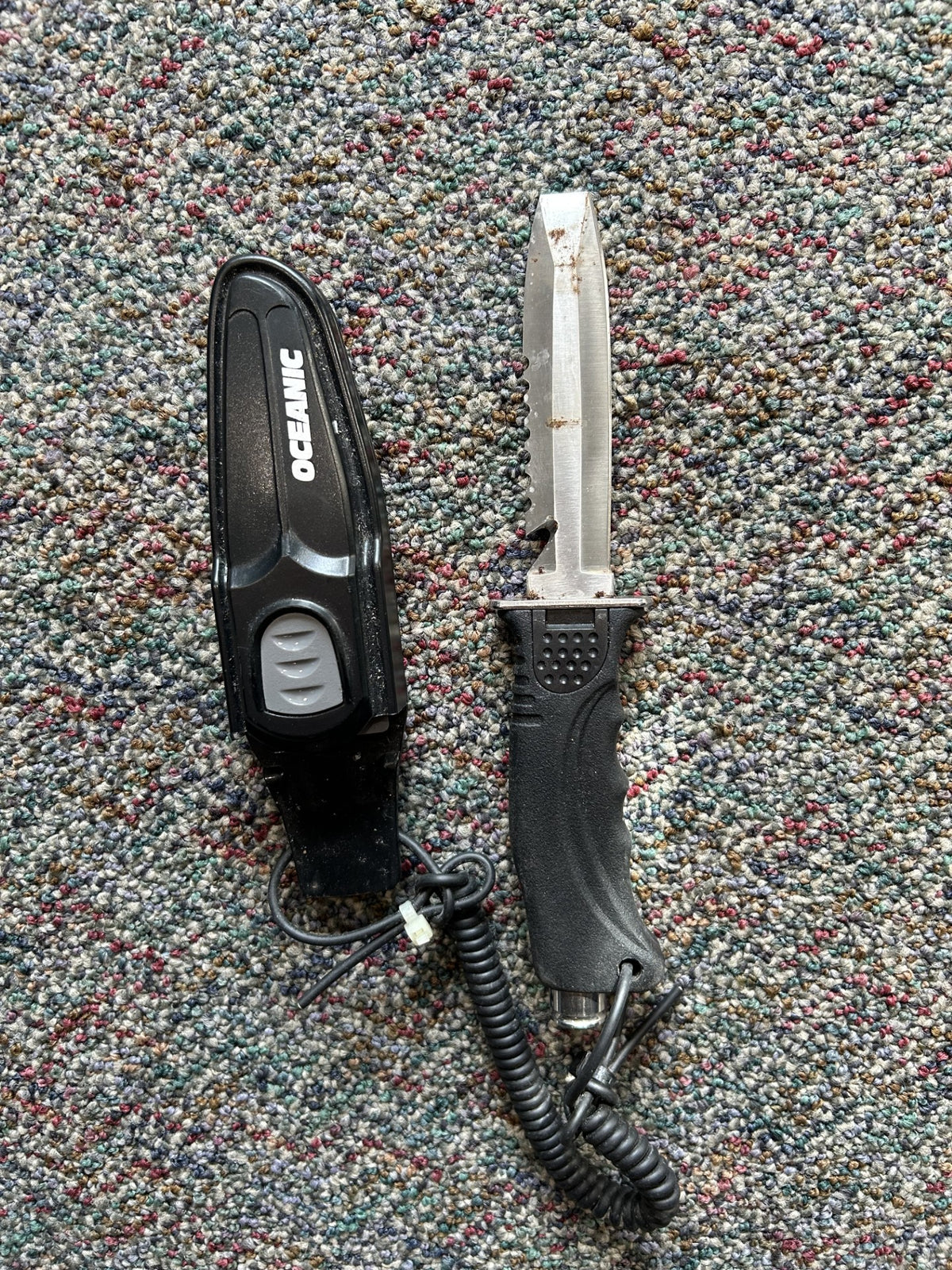 An OCEANIC brand black handheld device with a coiled cord and clip, shown next to a fixed-blade knife with a silver blade and black handle, both placed on a textured multicolor carpet.