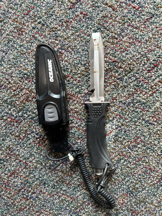 An OCEANIC brand black handheld device with a coiled cord and clip, shown next to a fixed-blade knife with a silver blade and black handle, both placed on a textured multicolor carpet.