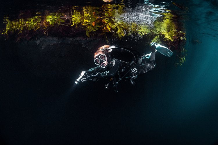 Scuba diver with a torch swimming beneath a weed-covered overhang in dark fresh water