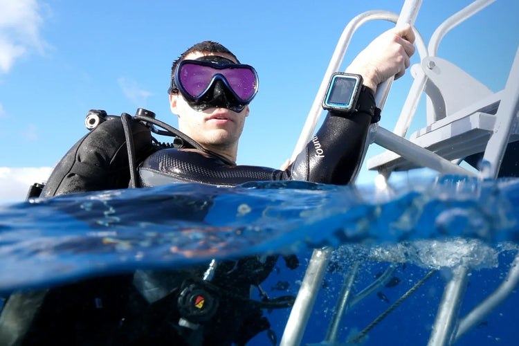 Scuba diver at the surface beside a boat ladder wearing a wrist-mounted dive computer