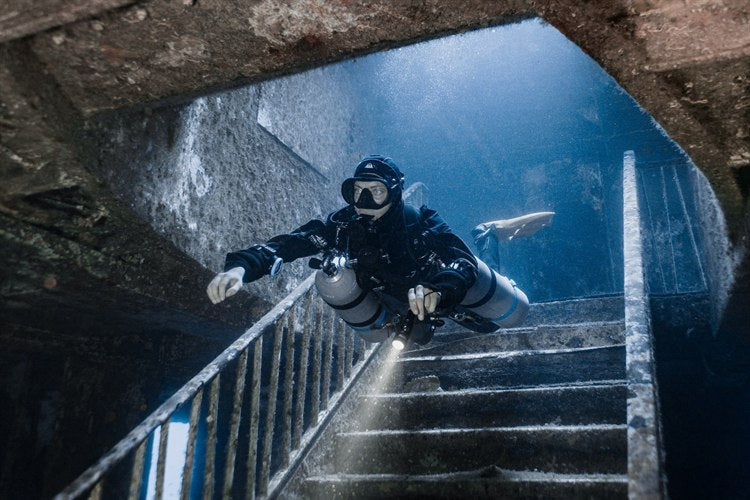 Diver in a hooded suit and sidemount cylinders finning down a stairwell inside a wreck